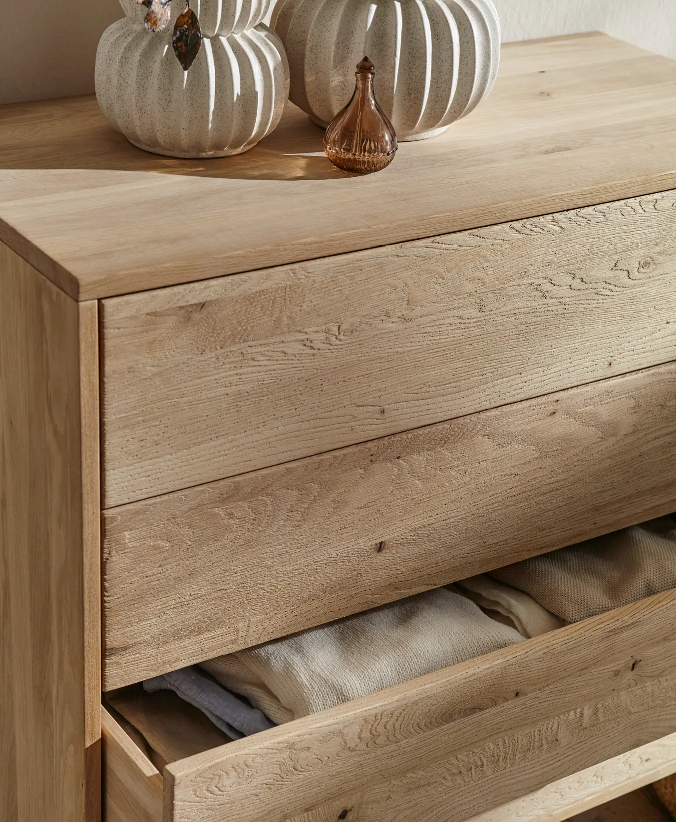 Detail einer Holzkommode mit geöffneter Schublade, Textilien im Inneren und Keramikvasen sowie Glasflasche oben